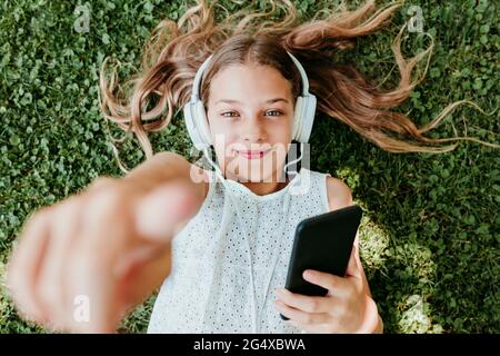 Smiling girl wearing headphones pointing while lying on grass Stock Photo