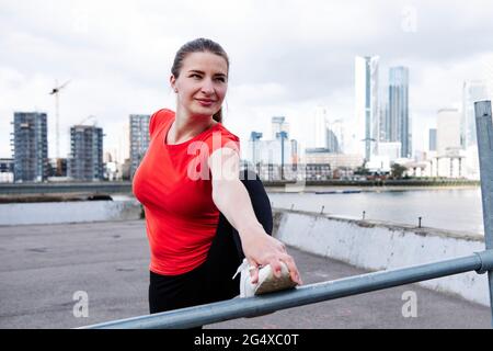 Smiling fit woman doing stretching exercise by river in city Stock Photo