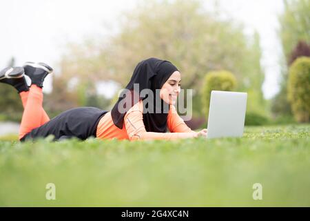 Young Arab woman lying on grass while using laptop in park Stock Photo