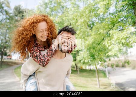 Cheerful boyfriend carrying girlfriend piggyback while covering eyes in park Stock Photo
