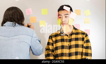 Confused young businessman with sticky notes on face standing by male colleague writing in office Stock Photo