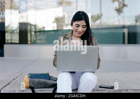 Smiling female freelancer using laptop while sitting on footpath Stock Photo