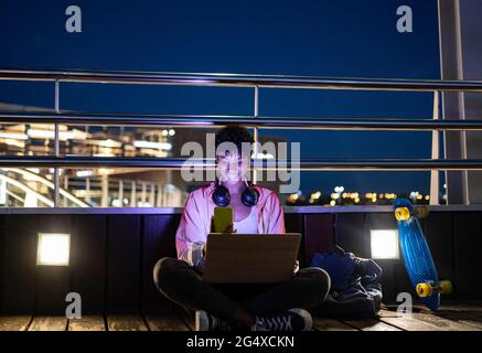 Young woman sitting cross-legged using smart phone by railing at night Stock Photo