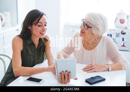 Senior woman talking with granddaughter using digital tablet while sitting at table Stock Photo