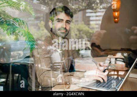 Businessman using laptop at table in restaurant seen through window glass Stock Photo
