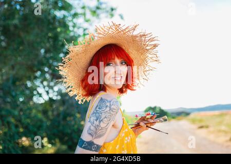 Smiling tattooed woman in straw hat holding paintbrushes and palette Stock Photo