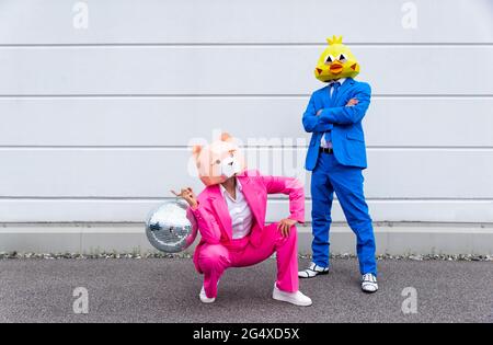 Man and woman wearing vibrant suits and animal masks posing with disco ball in front of white wall Stock Photo