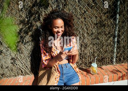 Smiling woman using smart phone while sitting on retaining wall Stock Photo