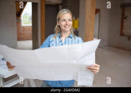 Smiling female architect looking away while holding blueprint at construction site Stock Photo