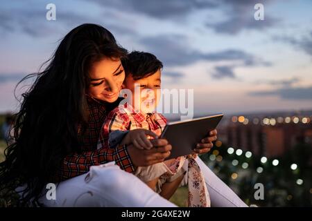 Happy son using digital tablet while sitting with mother during sunset Stock Photo
