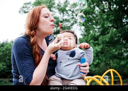 Mother blowing bubbles while holding son at park Stock Photo