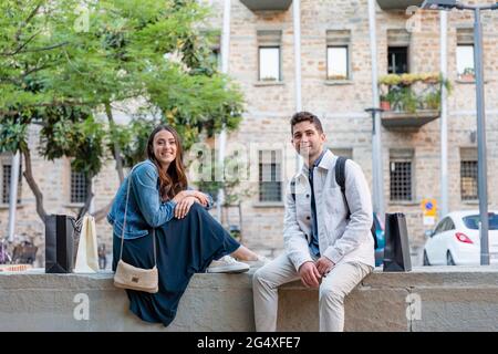 Smiling tourist couple sitting on concrete retaining wall Stock Photo