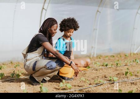 Female farmer teaching gardening to girl while crouching at greenhouse Stock Photo