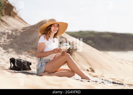 Smiling young woman wearing hat sitting with book on sunny day at beach during vacations Stock Photo