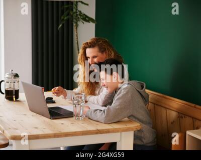 Smiling woman assisting son while e-learning on laptop at home Stock Photo