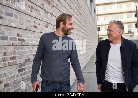Happy businessmen looking at each other while walking by wall Stock Photo