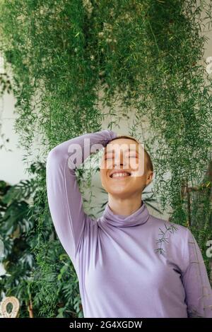 Happy woman with hand in hair and eyes closed by green plants Stock Photo