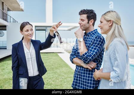 Female real estate agent showing house key to couple while standing at lawn