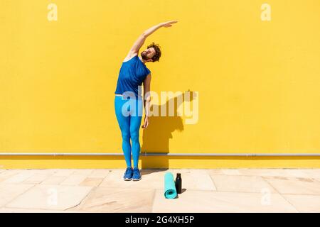 Man doing stretching exercise while standing in front of yellow wall Stock Photo