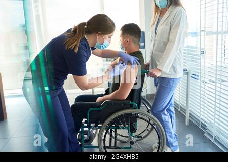 Female frontline worker giving COVID-19 vaccination dose to male patient sitting on wheelchair at center Stock Photo