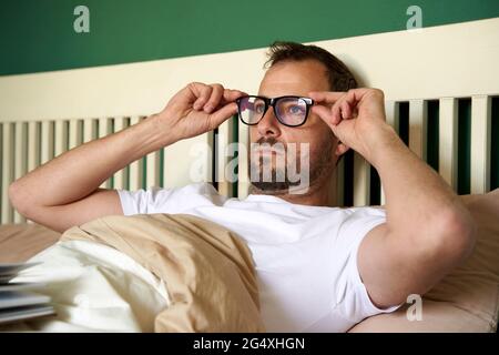 Man wearing eyeglasses while resting on bed in bedroom Stock Photo