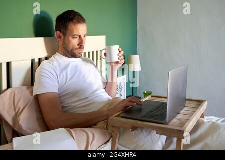 Mid adult man with coffee cup using laptop while resting in bedroom Stock Photo
