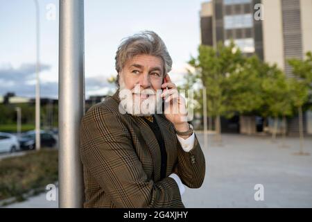Mature businessman leaning on pole while talking through mobile phone in city Stock Photo