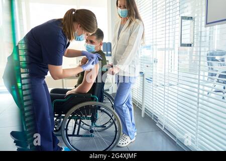 Female doctor giving COVID-19 vaccination dose to disabled man sitting on wheelchair at clinic Stock Photo