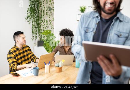 Male and female professionals with laptop discussing sitting at desk while colleague using digital tablet in office Stock Photo