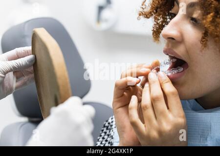 Female patient wearing dental aligner while looking at mirror held by dentist at clinic Stock Photo