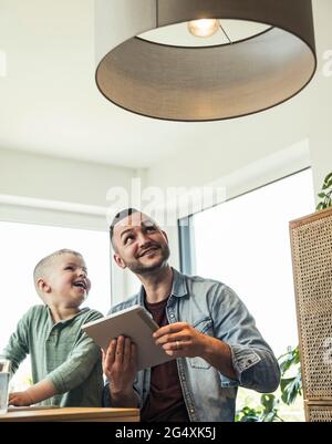Smiling son sitting with father controlling pendant light through tablet at smart home Stock Photo
