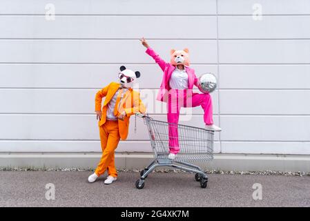 Man and woman wearing vibrant suits and animal masksÂ posing with shopping cart and disco ball in front of white wall Stock Photo