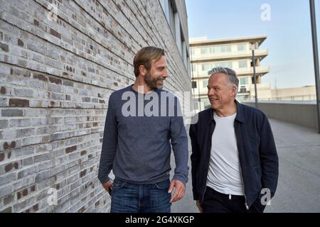 Happy mature businessmen talking while looking at each other walking by wall Stock Photo