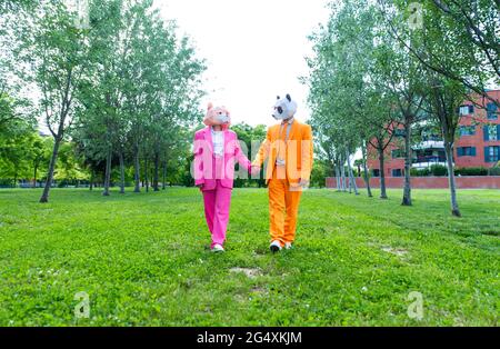 Adult couple wearing vibrant suits and animal masks holding hands while walking together in green park Stock Photo
