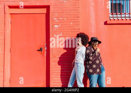 Smiling female friends standing back to back by red building Stock Photo