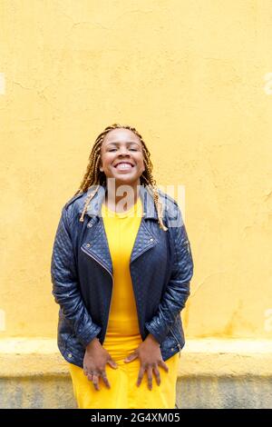 Smiling woman with braids standing in front of yellow wall Stock Photo