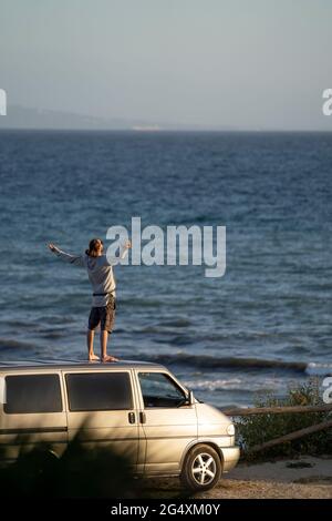 Young man with arms outstretched standing on van roof by sea Stock Photo