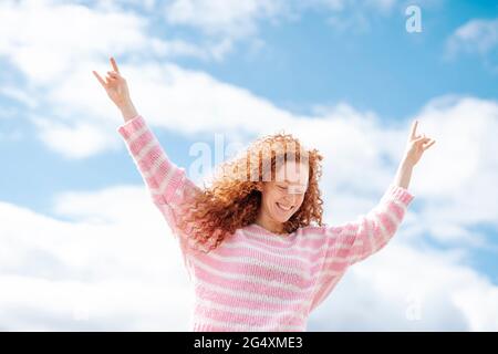 Cheerful young curly haired woman with arms raised standing under sky Stock Photo