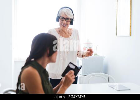 Smiling senior woman listening music through headphones while looking at granddaughter using smart phone Stock Photo