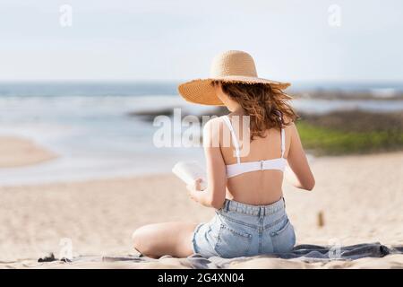 Woman wearing sun hat reading book while sitting at beach during vacations Stock Photo