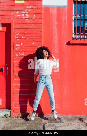 Cheerful woman jumping in front of red building during sunny day Stock Photo