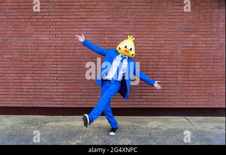 Man wearing vibrant blue suit and bird mask posing on one leg in front of brick wall