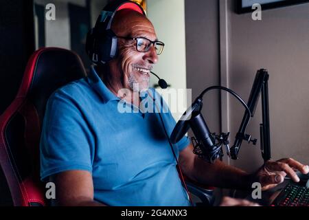 Smiling senior man wearing headphones using keyboard while playing video game at home Stock Photo