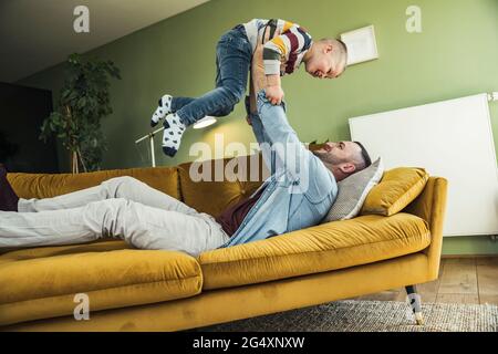 Playful father lifting up son while lying on sofa in living room at home Stock Photo