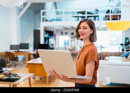 Happy female professional using laptop while standing in creative office Stock Photo