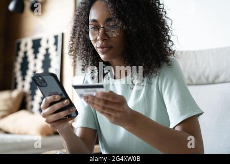 Happy millennial mixed race woman holding banking credit card, entering payment information in smartphone application Stock Photo