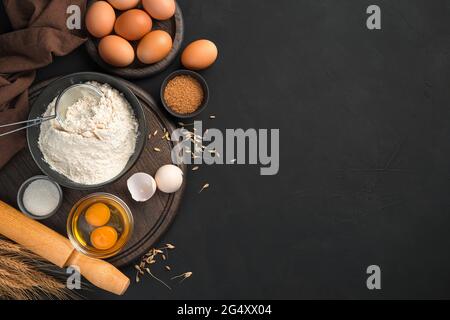 Pastries, ingredients for making a dish of dough on a black background. Flour, eggs, sugar and rolling pin. Stock Photo