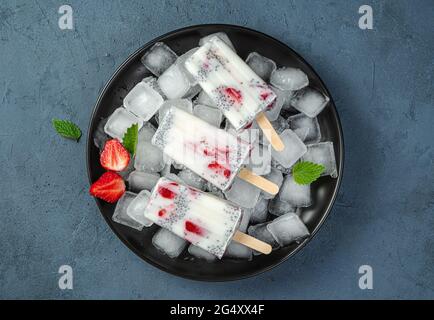 Three ice creams with strawberries and chia seeds on ice cubes in a black plate on a blue background Stock Photo