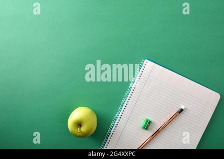 Open spiral notebooks with pencil and sharpener on top and green apple aside on green background with space for text. Top view. Stock Photo