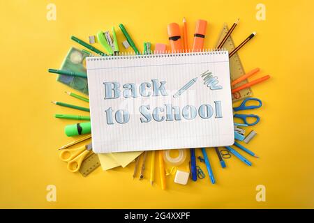 Educational notebook with blue lines open on pile of school supplies with text back to school on yellow background. Top view. Stock Photo